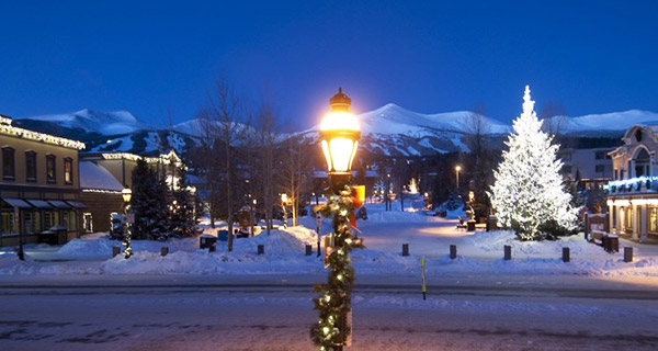Holiday lights on trees, buildings and light posts in downtown Breckenridge with the mountains in the background.