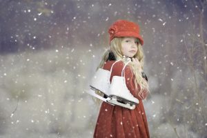 A young girl carrying ice skates with snow flakes falling around her.